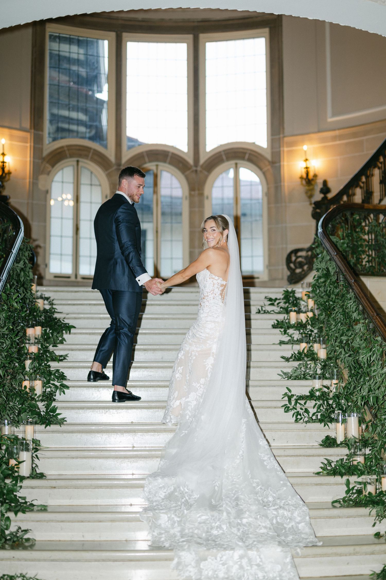 Armour House Wedding - Bride and groom hold hands on elegant staircase adorned with greenery and candles in a grand wedding venue.
