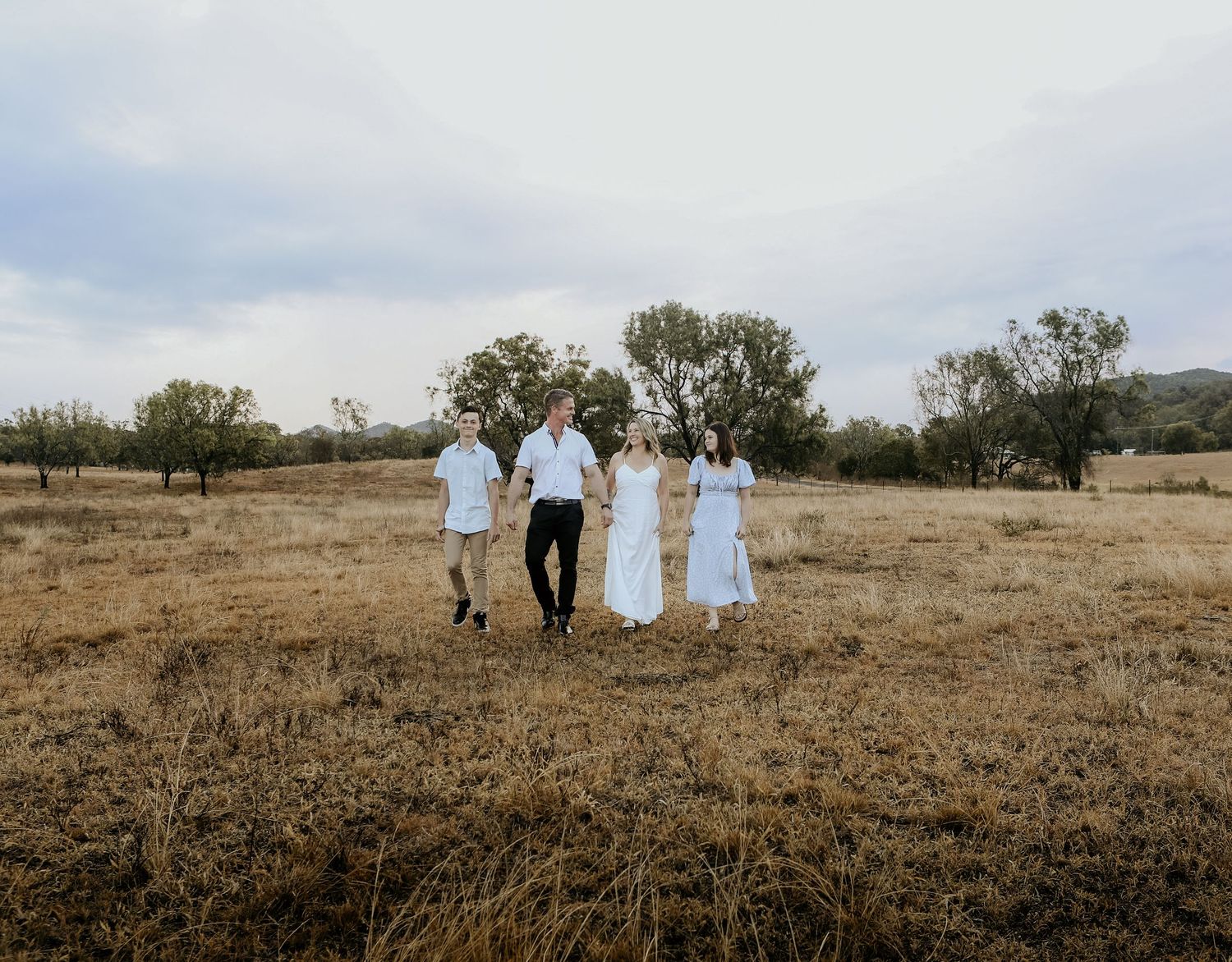 Four people in white attire walk through a golden dry field with trees under an overcast sky.