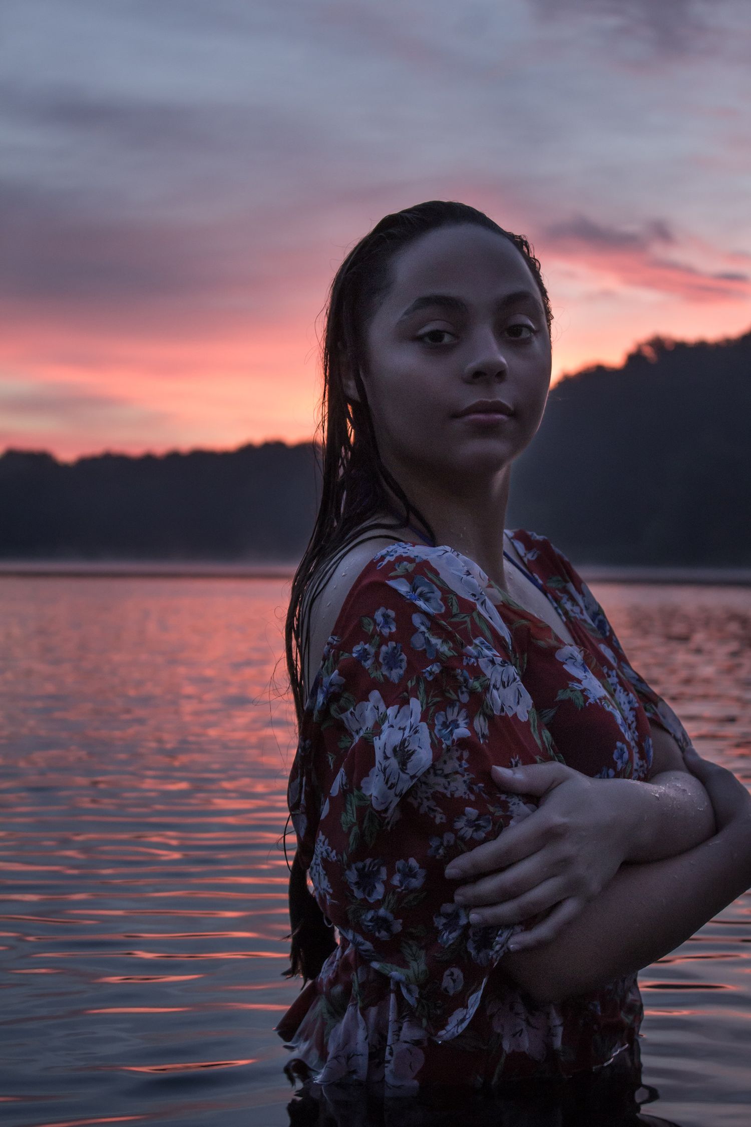 Woman in floral dress standing in lake at dramatic pink sunset with wet hair crossed arms.