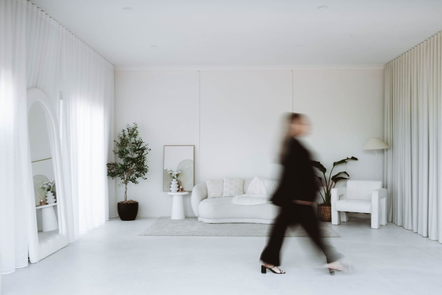 Blurred motion of a businesswoman walking through a modern white minimalist living room with sofa and plants.