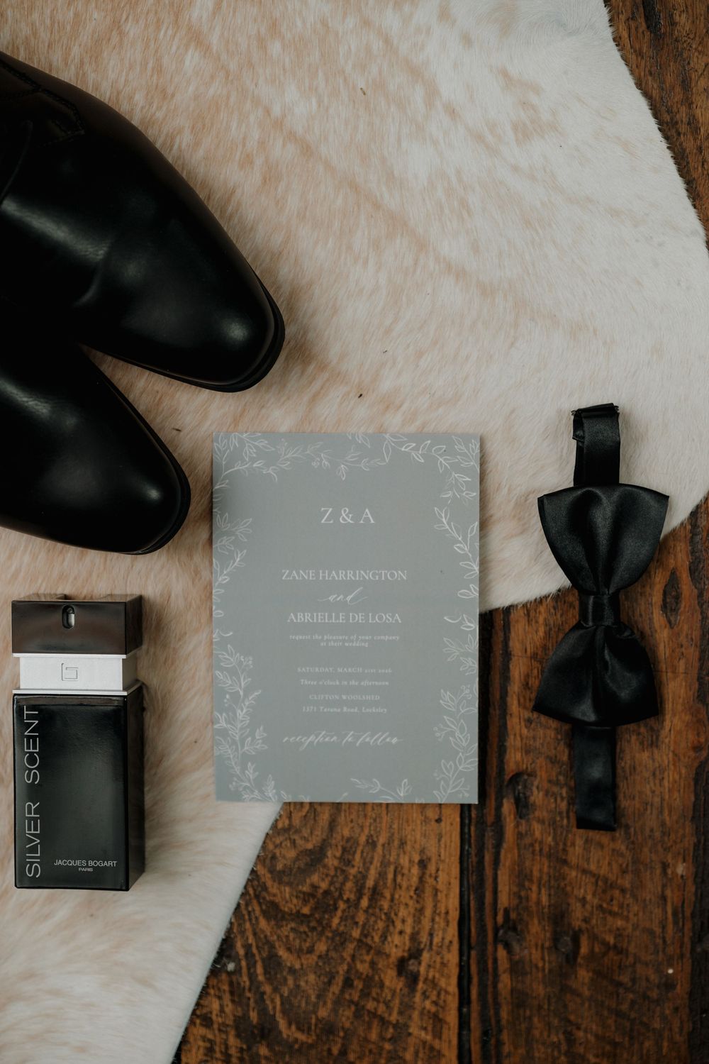 Elegant groom flat lay with gray wedding invitation, black bow tie, dress shoes, and Silver Scent cologne on a fur rug.