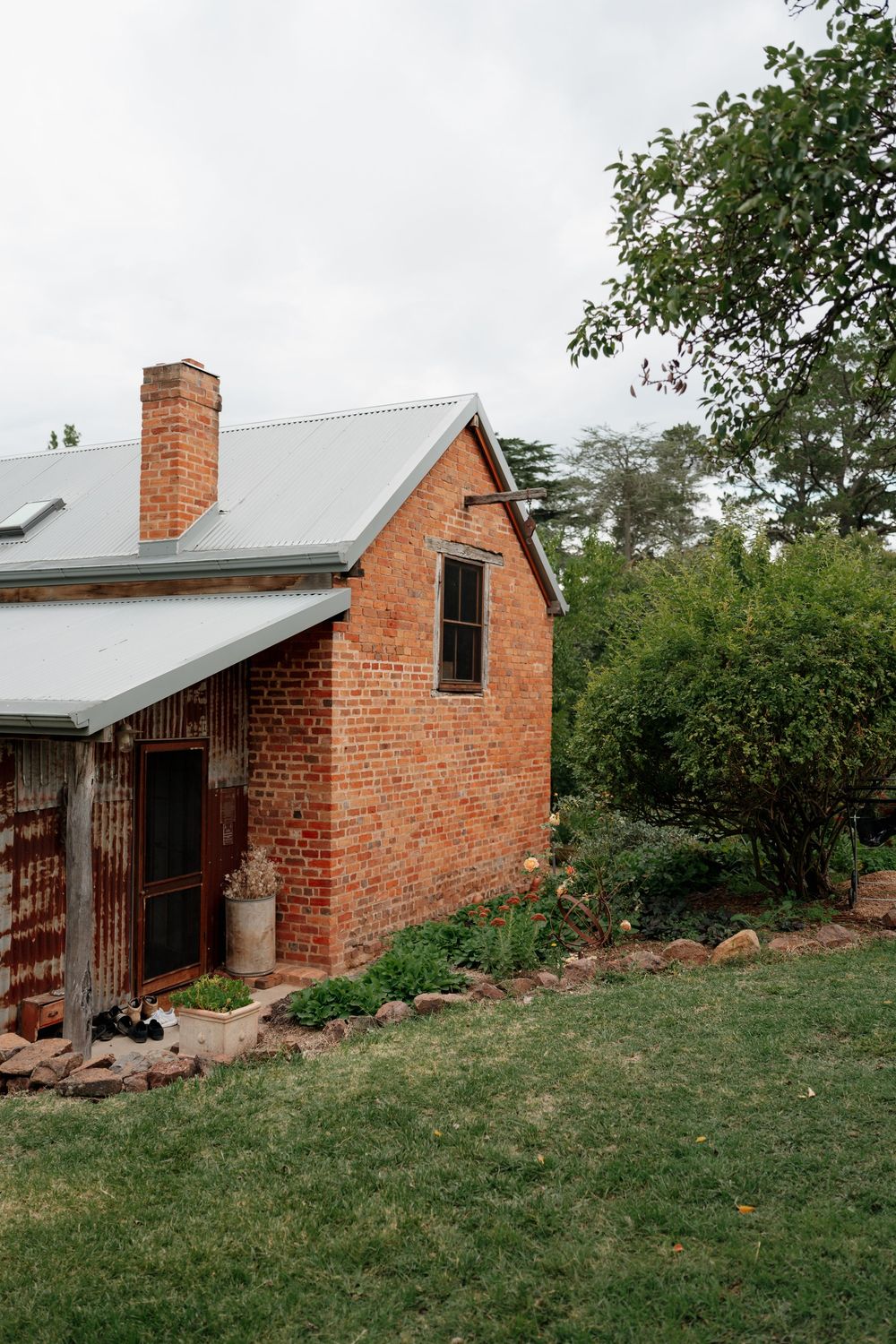 Rustic red brick cottage with metal roof and chimney surrounded by lush green garden trees.