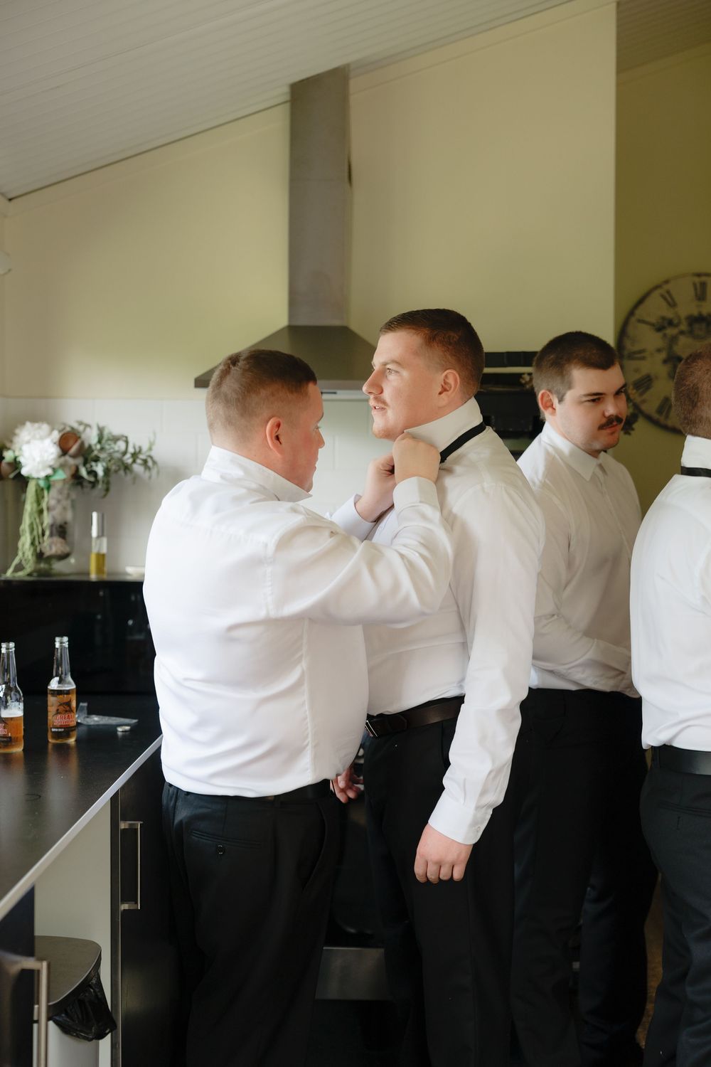 Groomsmen helping groom adjust black bow tie while getting ready in modern kitchen.