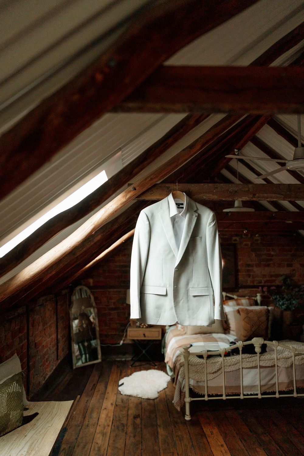 White wedding suit jacket hanging in rustic attic bedroom with wooden beams and brick walls.