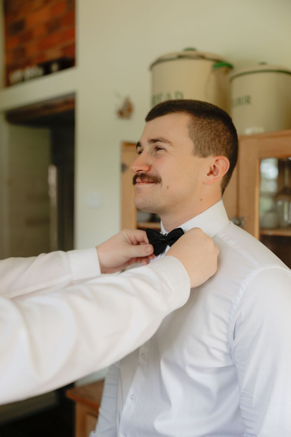Groom in white shirt getting black bow tie adjusted by helper during wedding preparations.
