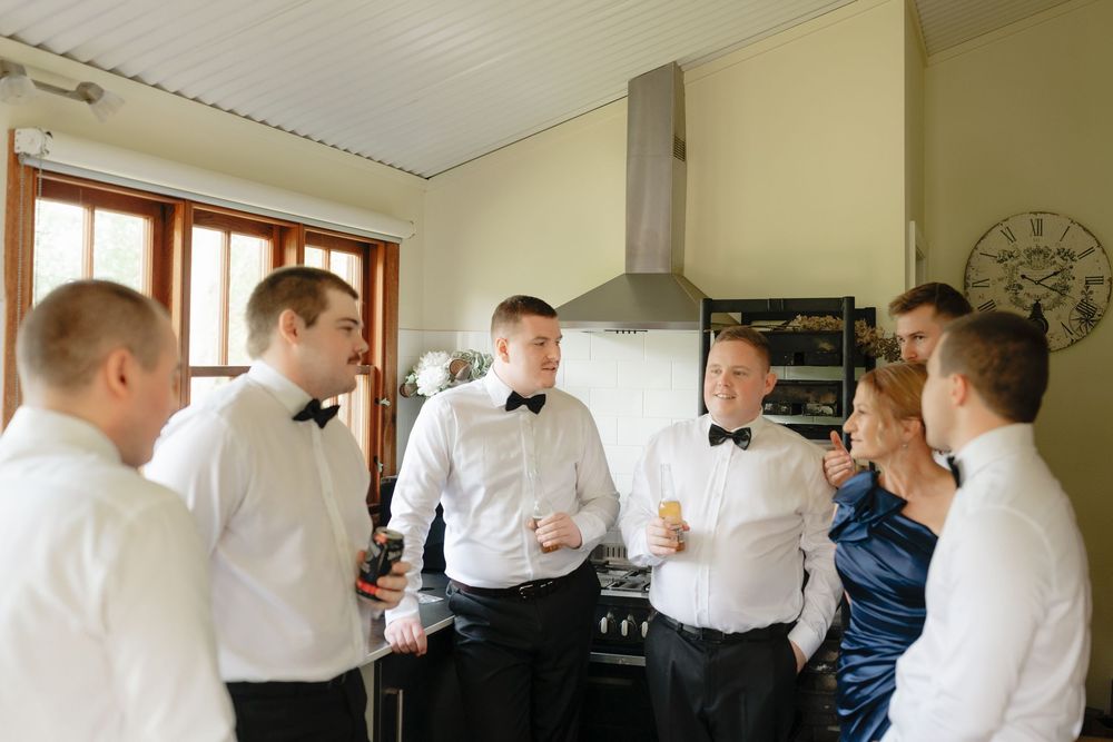 Groomsmen in white shirts and bow ties socializing in a kitchen before a wedding ceremony.