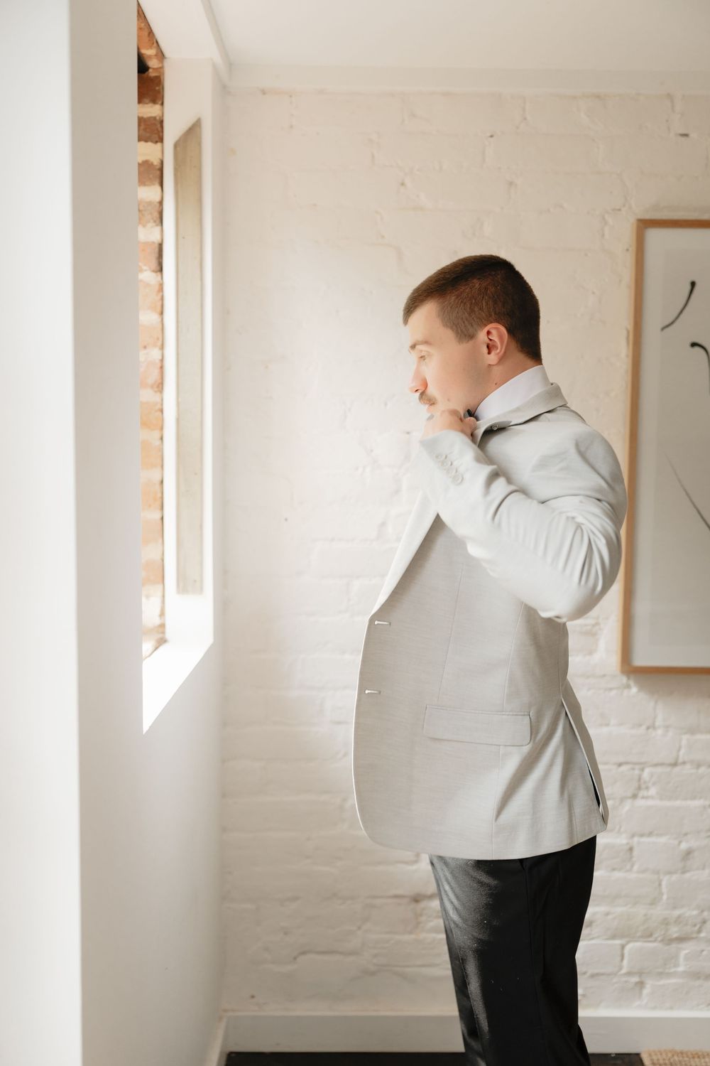 Groom in a light gray suit jacket adjusting his bow tie while standing by a window in a white brick room.