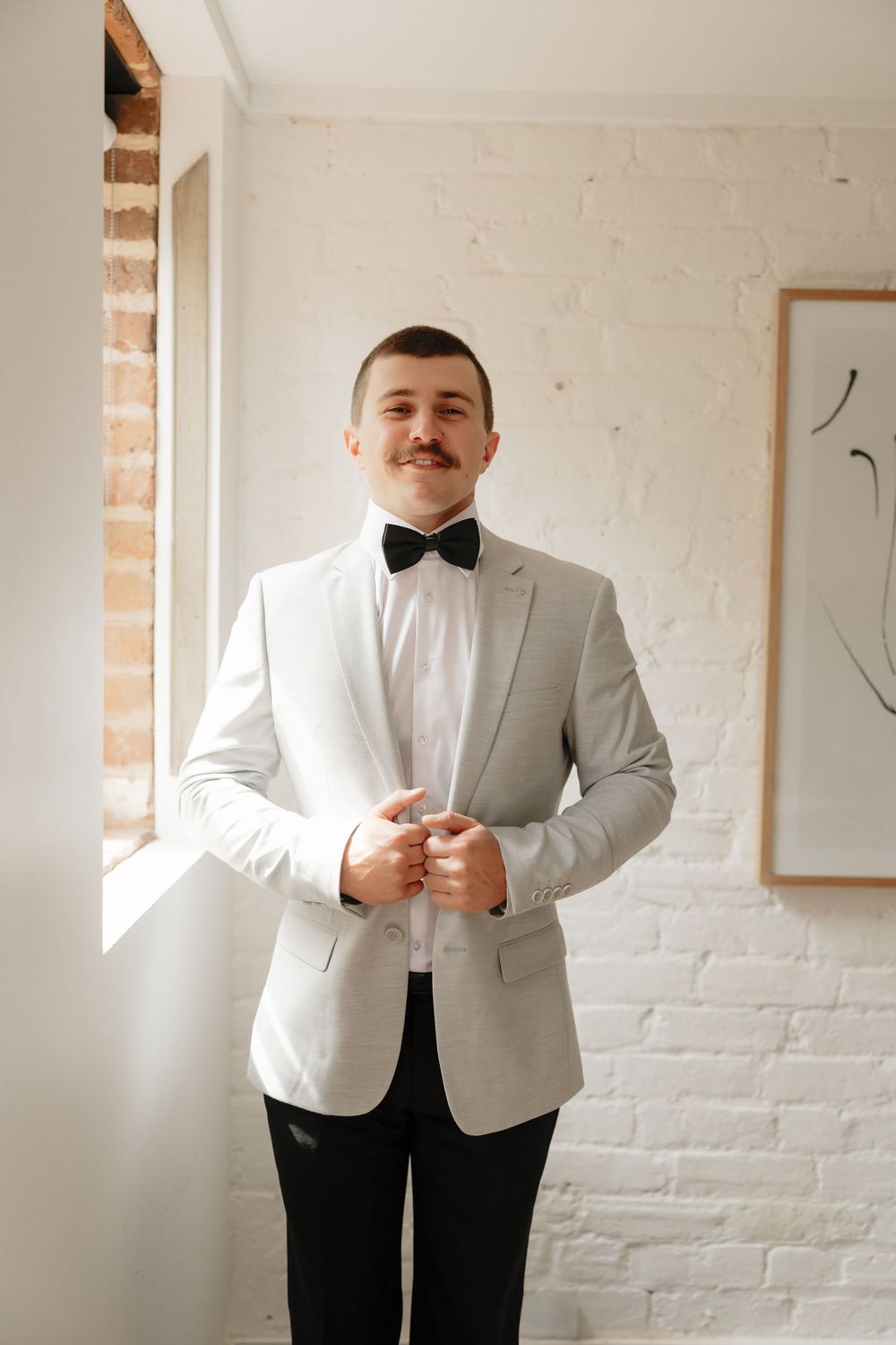 Groom in light gray suit and black bow tie buttoning jacket indoors before wedding ceremony.