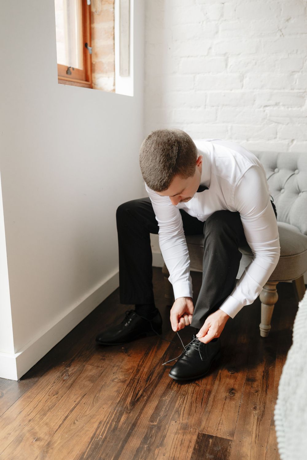 Groom in white dress shirt and black trousers tying his black leather dress shoes while seated on a tufted chair.