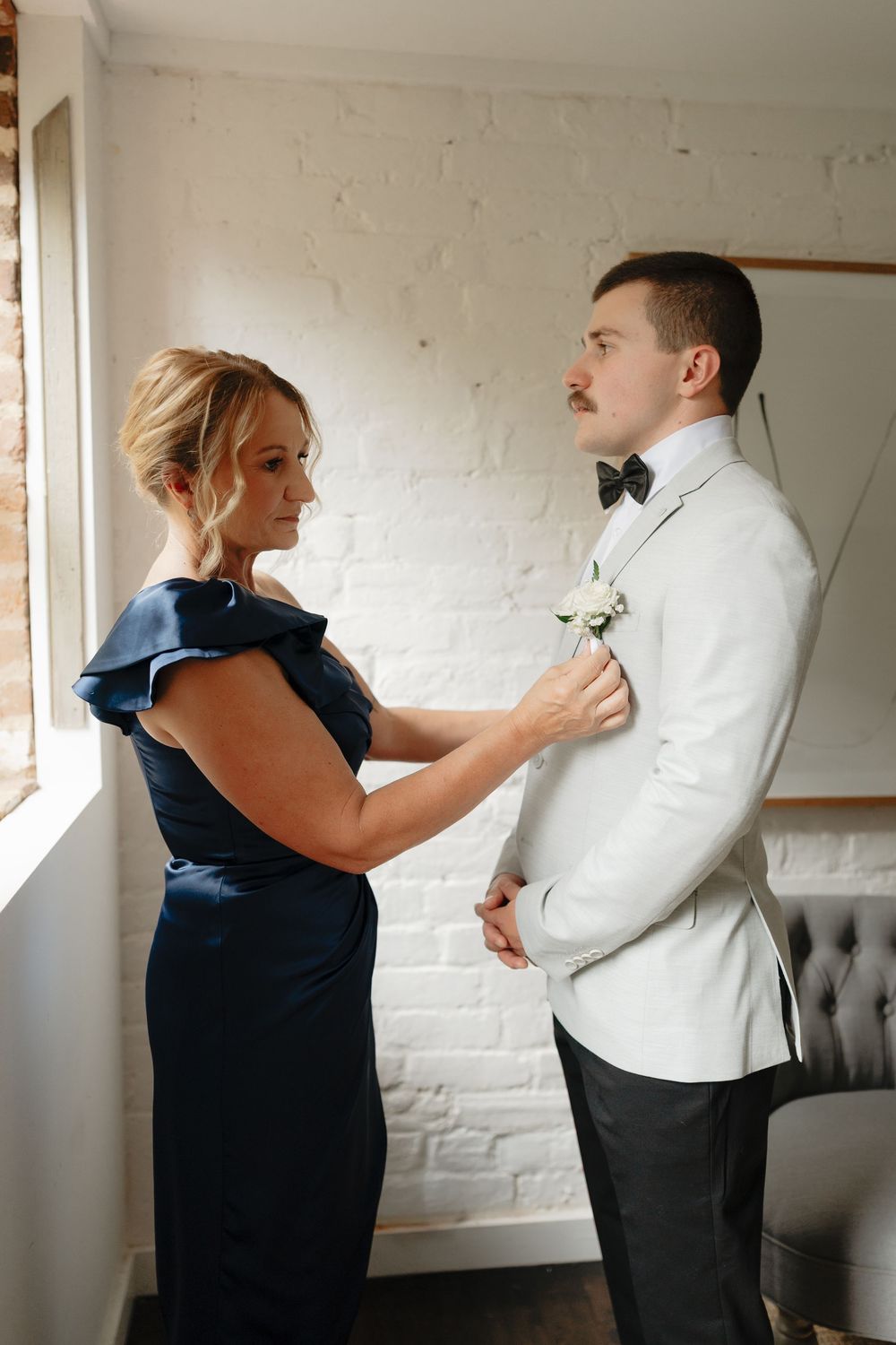 Woman in navy blue dress pinning boutonniere on groom in white tuxedo jacket indoors.