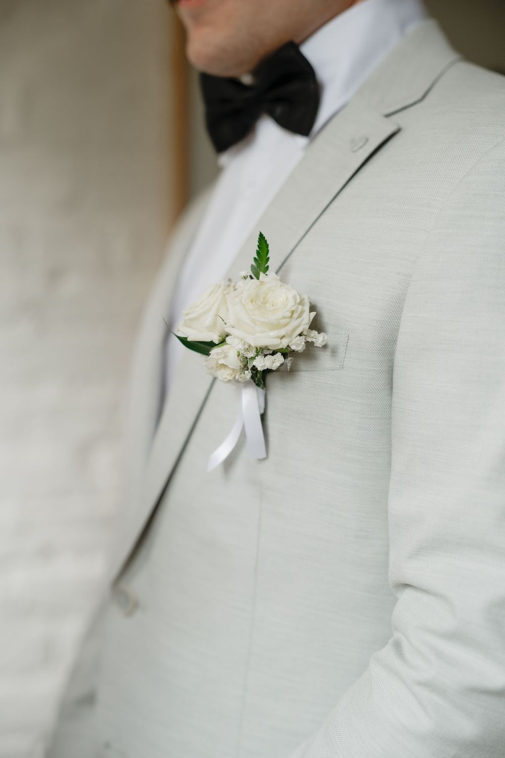 Close-up of groom in light gray suit with black bow tie and white rose boutonniere with greenery accent.