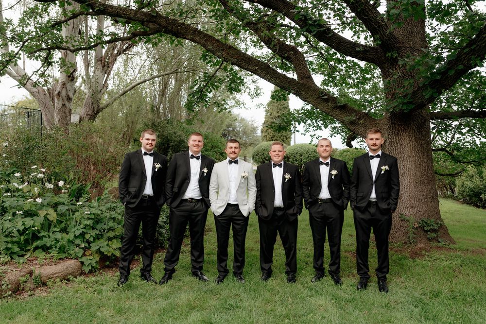 Six groomsmen in black tuxedos pose outdoors under a large tree, with one in a white jacket standing center.