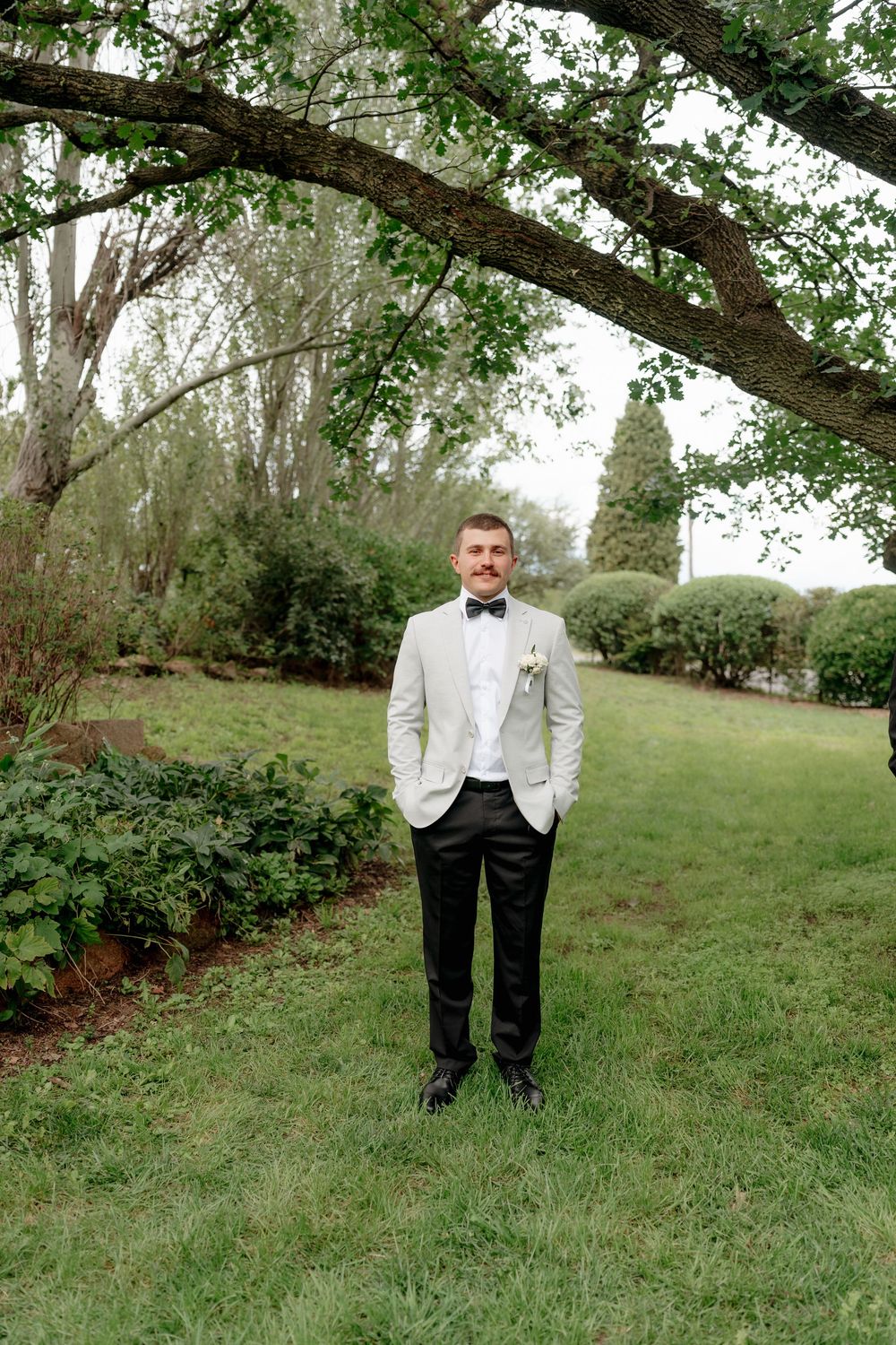 Groom in white tuxedo jacket and black bow tie standing in lush garden under tree branches.