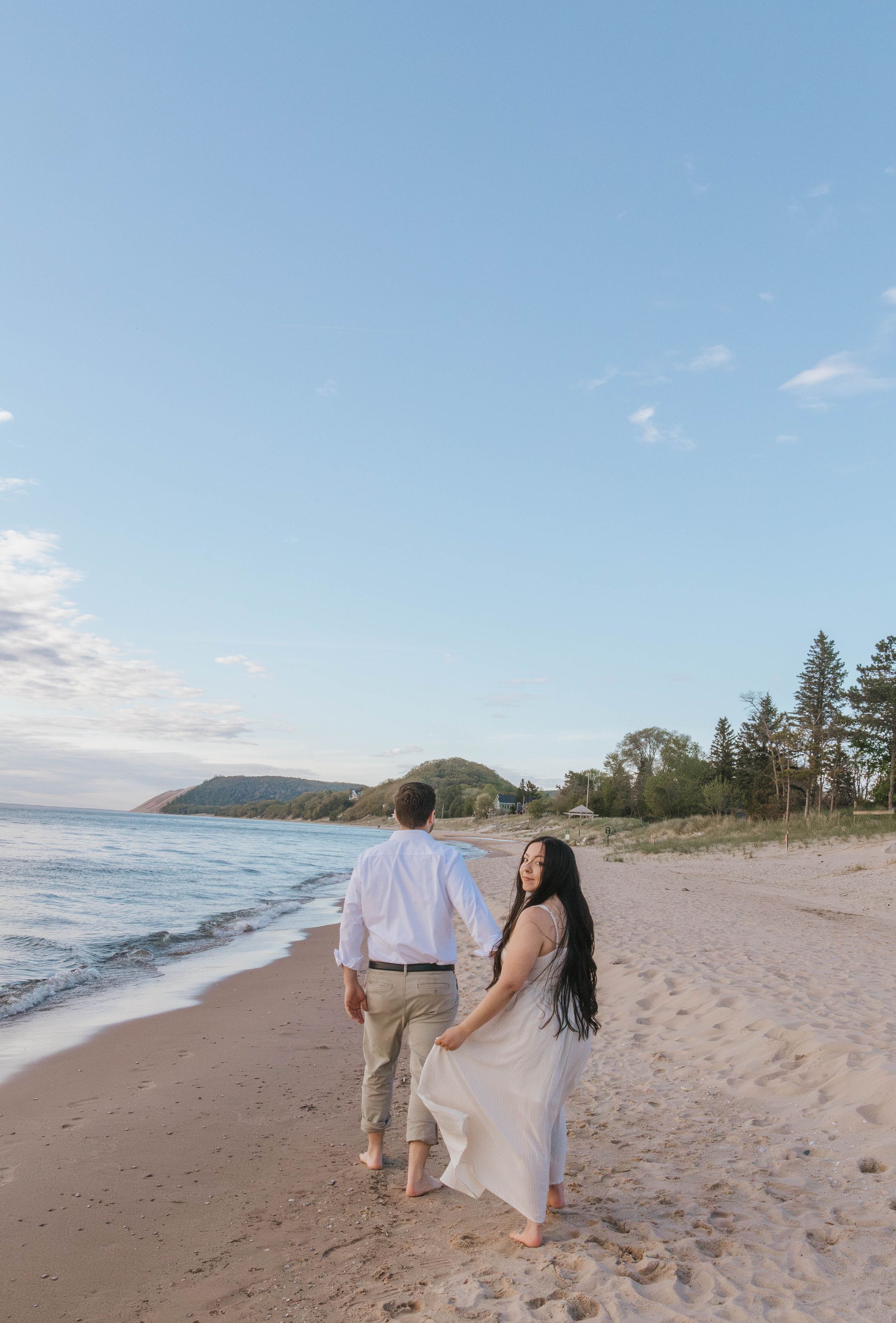 Couple walking hand in hand on a sandy beach at golden hour, woman in white dress looks back, trees and bluff in background.