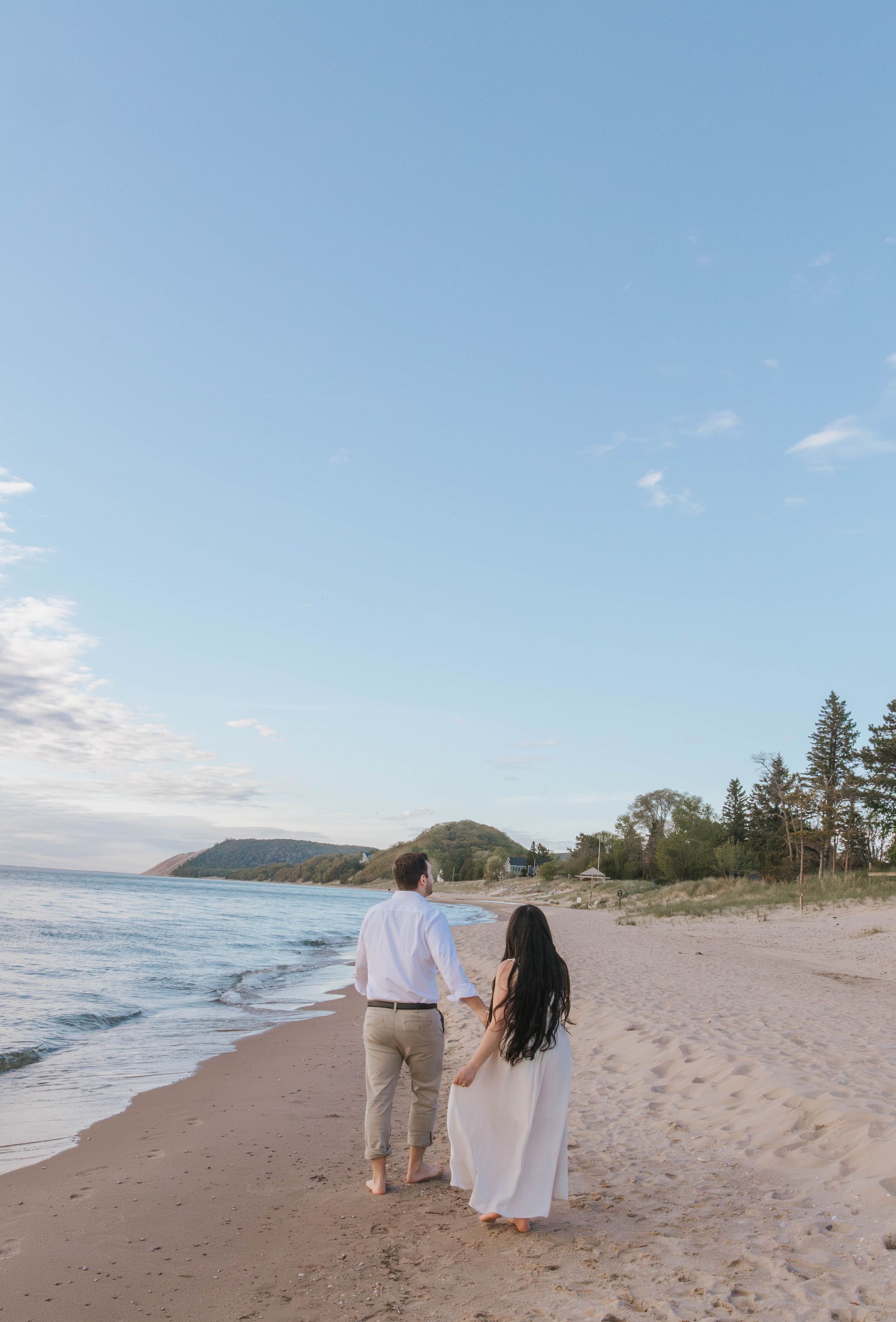 A couple walks hand-in-hand along a sandy beach at sunset, with trees and a hill in the background.