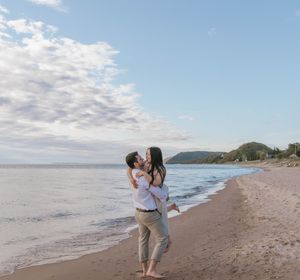 Couple embracing and kissing on a sandy beach at dusk with calm ocean waves and green hills in background.