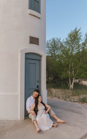 Couple sitting together against a white lighthouse with a blue door, surrounded by sand dunes and trees at golden hour.
