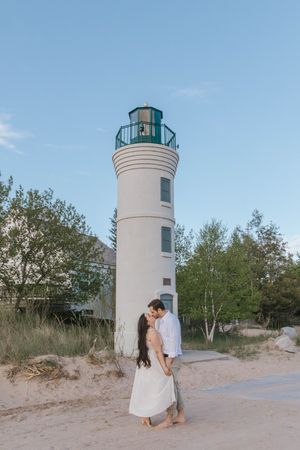 Couple sharing a romantic moment in front of a white lighthouse on a sandy beach surrounded by green trees.