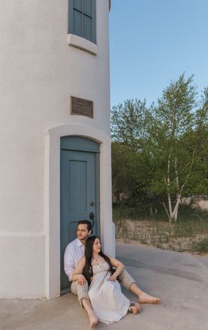 Couple sitting together at base of white lighthouse with teal door, sandy dunes and trees in background.
