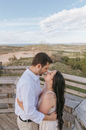 Couple sharing a romantic almost-kiss on a wooden deck overlooking sandy dunes and ocean under a blue sky.