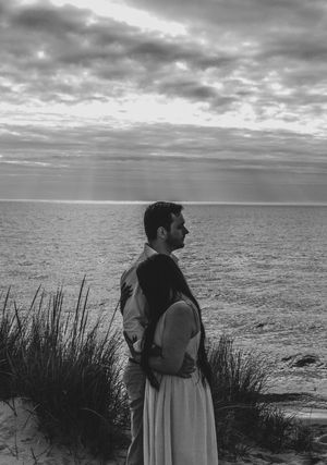Black and white photo of a couple embracing on a sandy beach at sunset with dramatic cloudy sky.