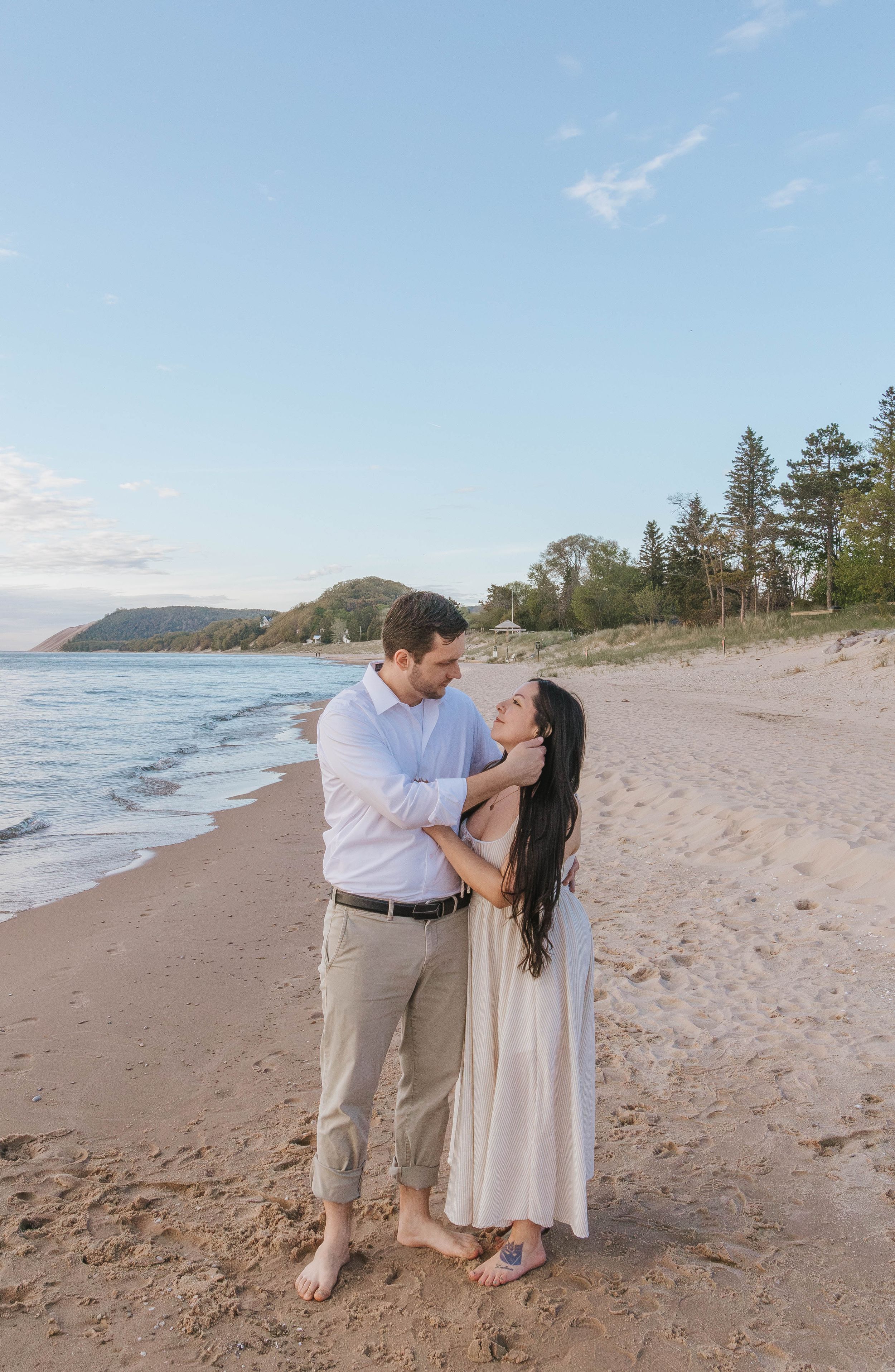 Couple embracing on a sandy beach at sunset, woman in white dress, man in light blue shirt, with trees and calm water behind.
