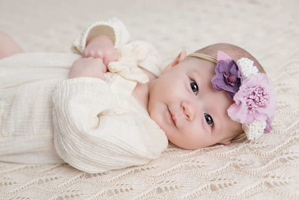 Adorable baby girl in white knit outfit with purple floral headband lying on a soft cream blanket.