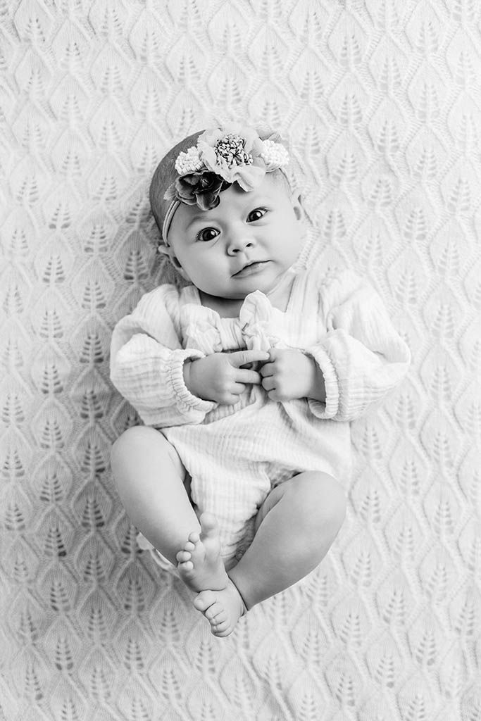 Black and white photo of an adorable baby girl wearing a floral headband, lying on a patterned blanket.