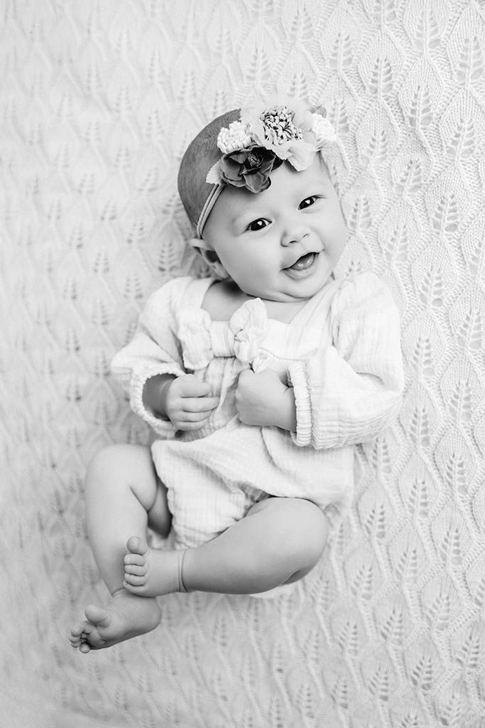 Smiling baby girl in white knit outfit and floral headband lying on textured white blanket, black and white photo.