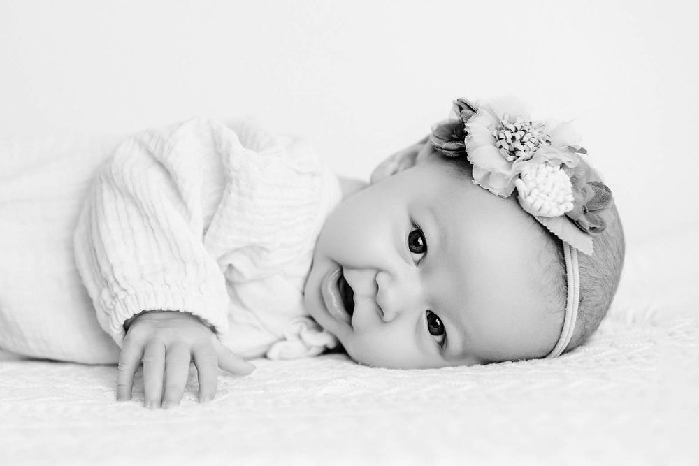 Black and white portrait of a smiling baby girl wearing a floral headband, lying on a soft white blanket.