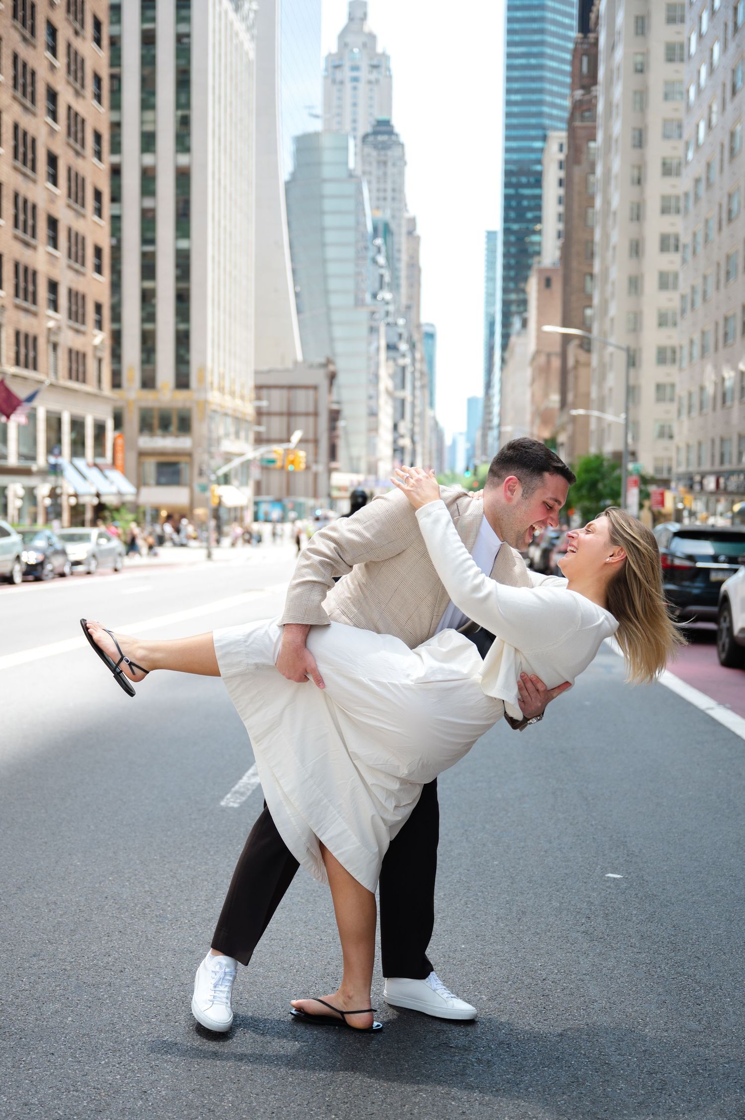 Couple dancing in dip pose on NYC street, woman in white dress, man in beige blazer, skyscrapers in background after their proposal.