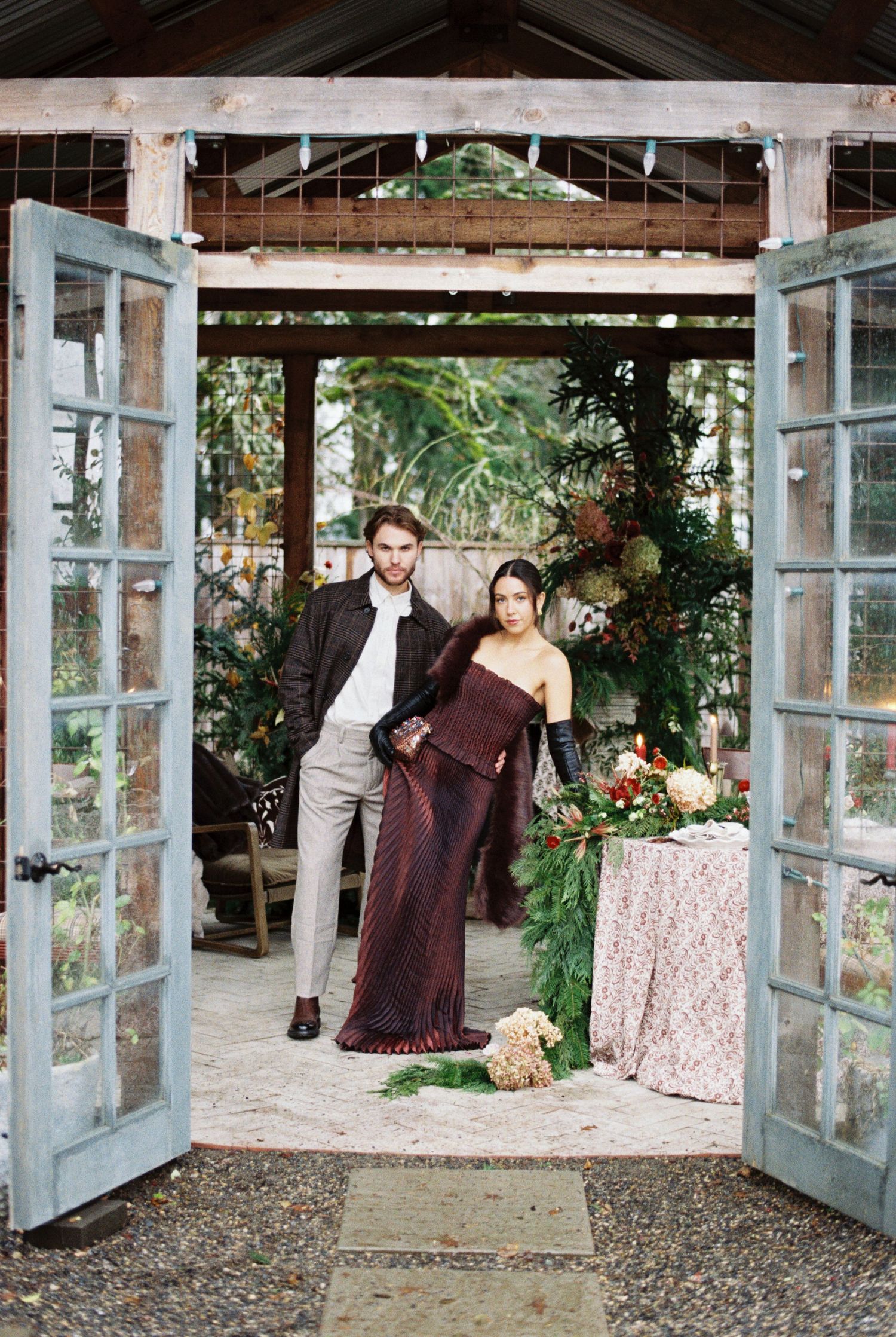 Couple poses in greenhouse doorway, woman in burgundy sari, man in dark jacket, surrounded by lush greenery and floral arrangements.