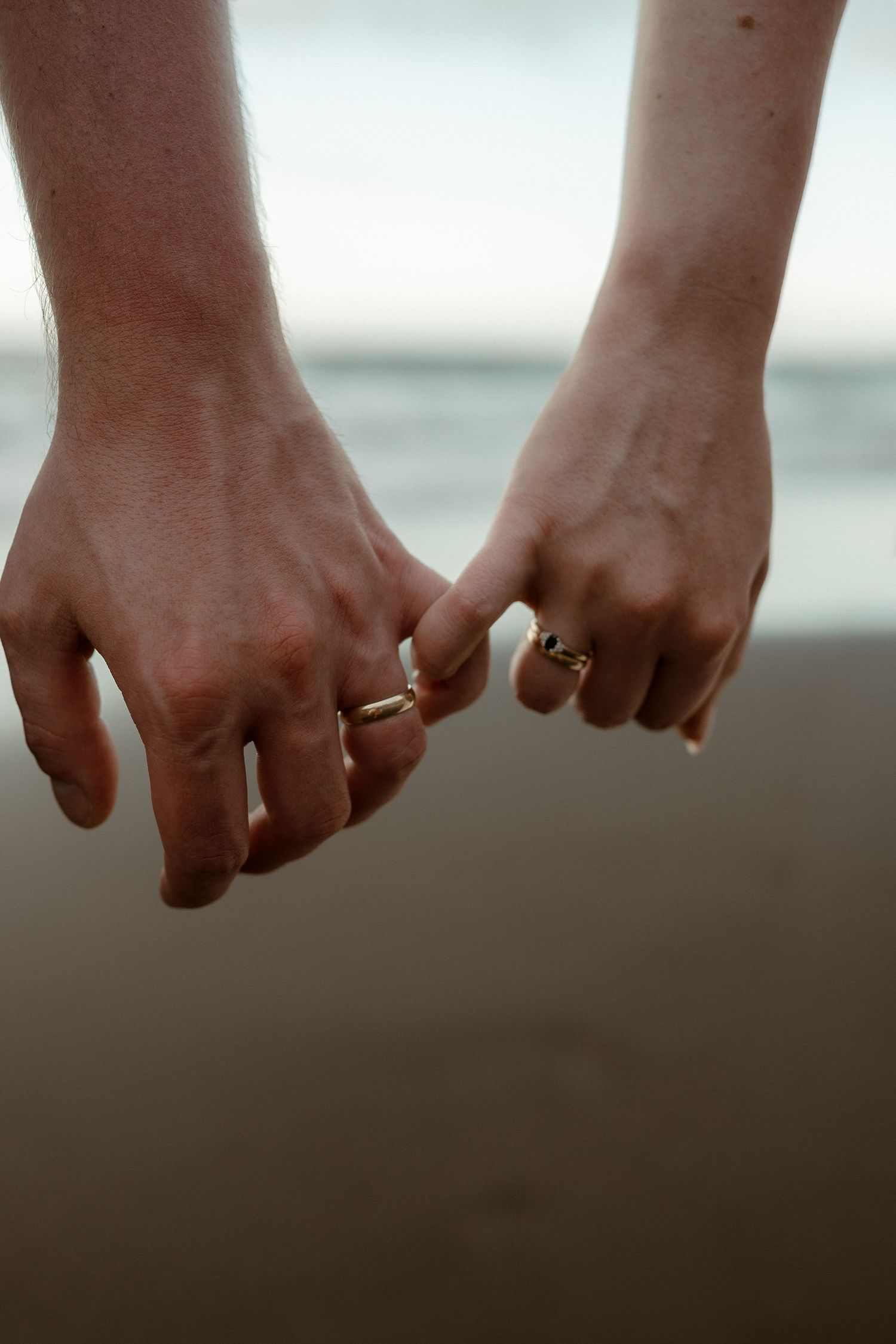 Two people holding pinkies together on a beach, one wearing a silver bracelet and ring, symbolizing love and connection.