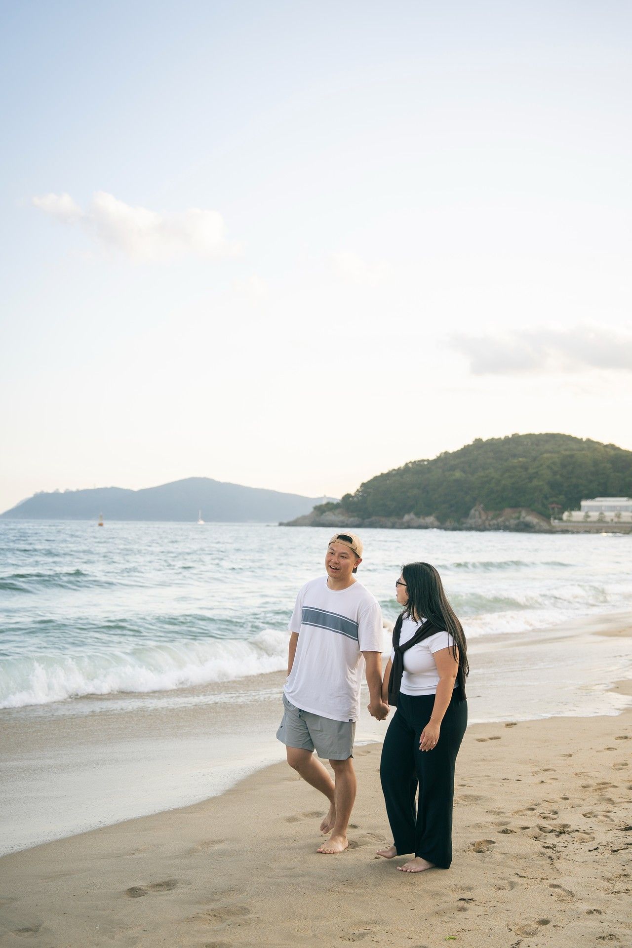Couple holding hands and walking barefoot on a sandy beach at sunset with ocean waves and green hills in background.