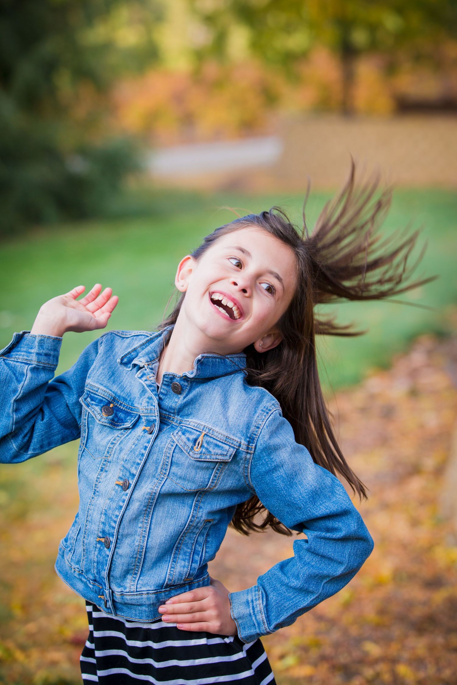 Young girl in denim jacket laughing joyfully outdoors, long hair flying in autumn park setting.