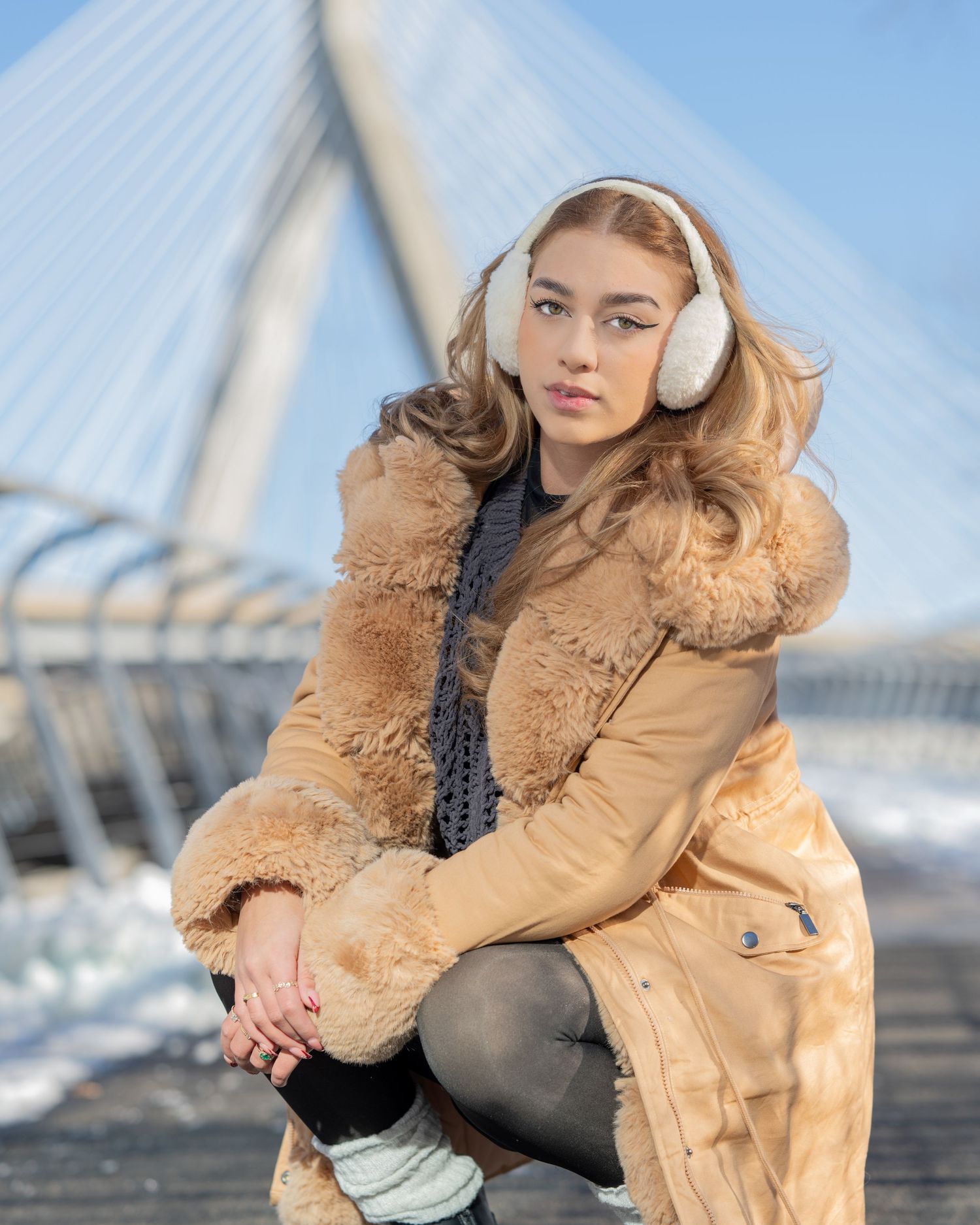 Young woman in a tan fur-trimmed coat wearing white earmuffs on a bridge in winter sunshine posed for portrait photo in Boston, MA