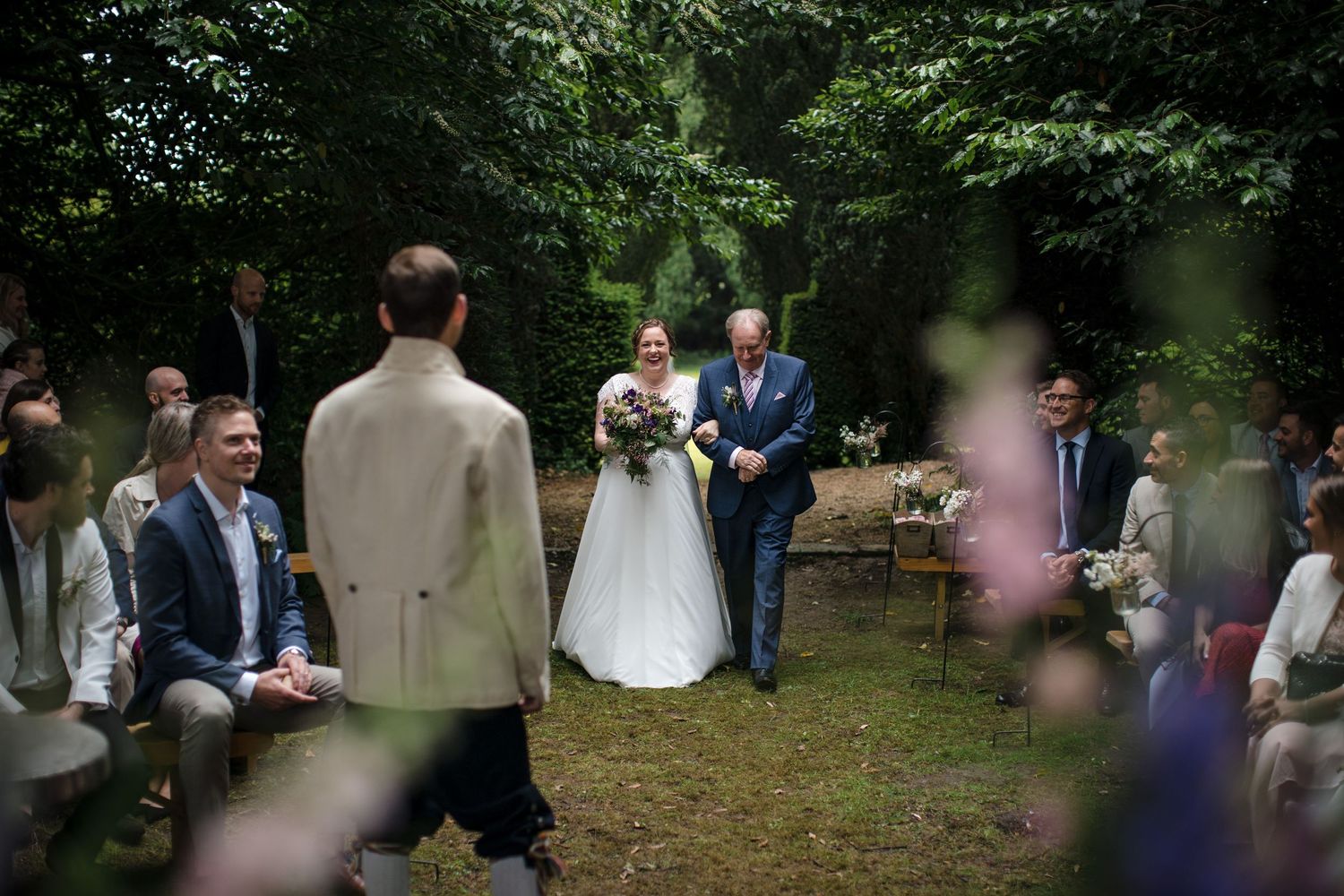 Bride walks down outdoor aisle with father at garden wedding ceremony, guests seated among lush green trees.