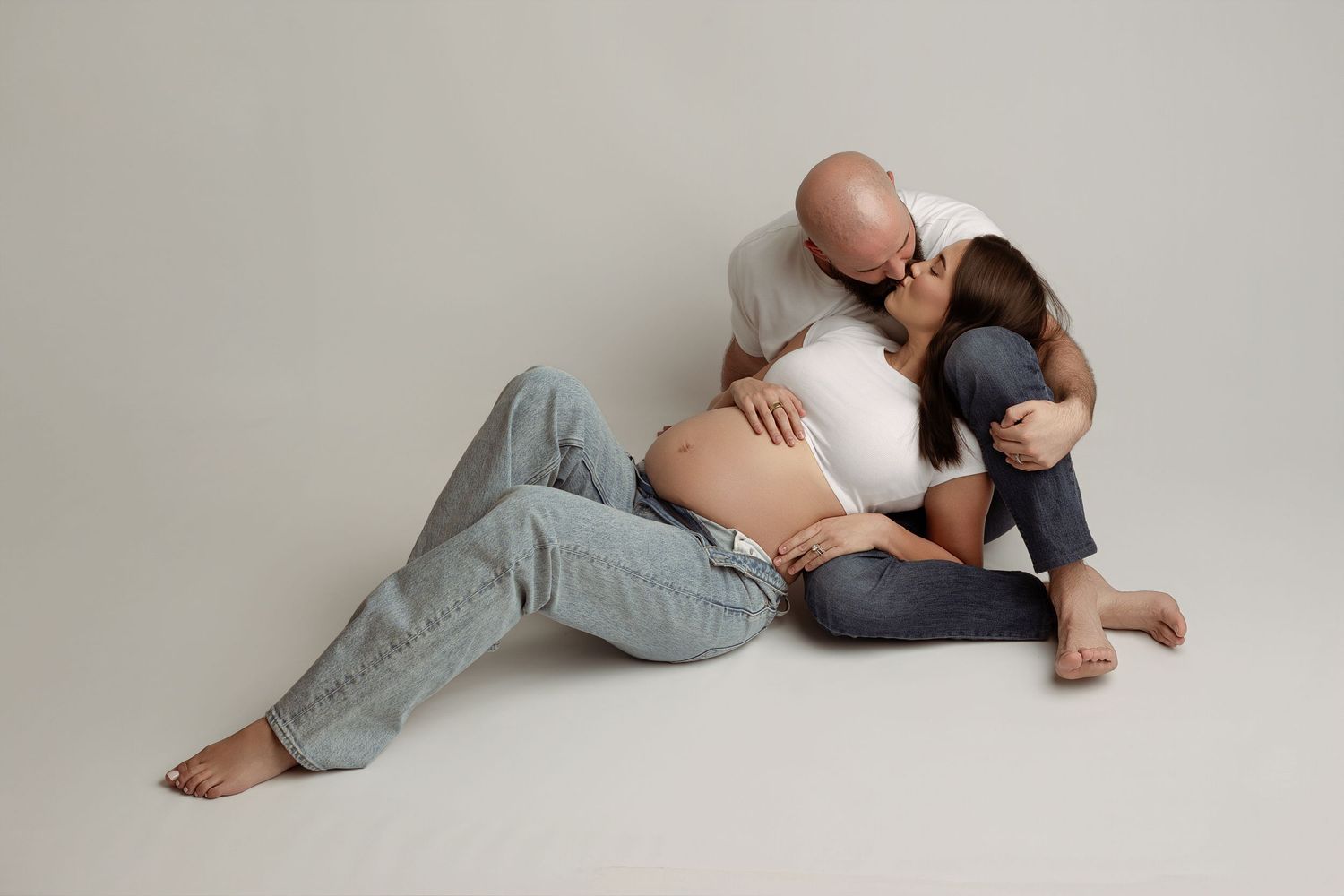Expecting couple sitting on floor, man kissing woman while gently touching her pregnant belly.