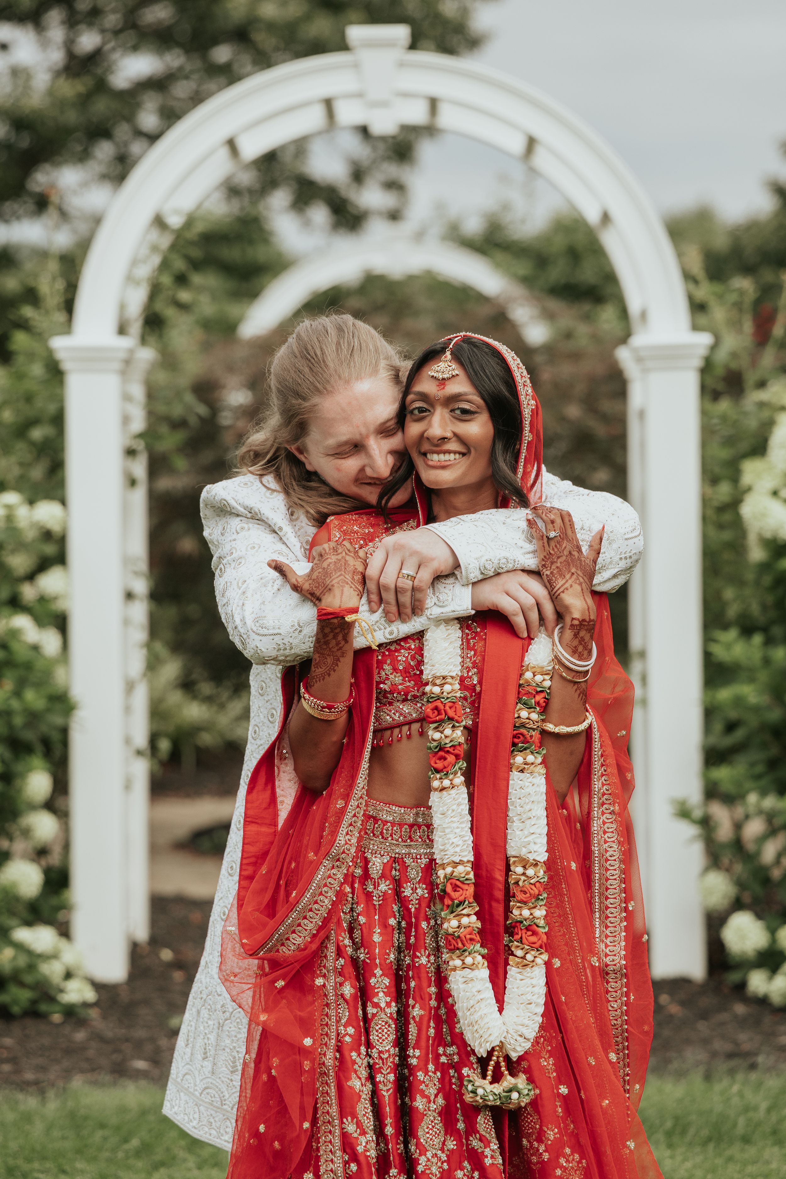 Groom in white sherwani embracing bride in red lehenga with mehndi and floral garland at outdoor wedding arch.