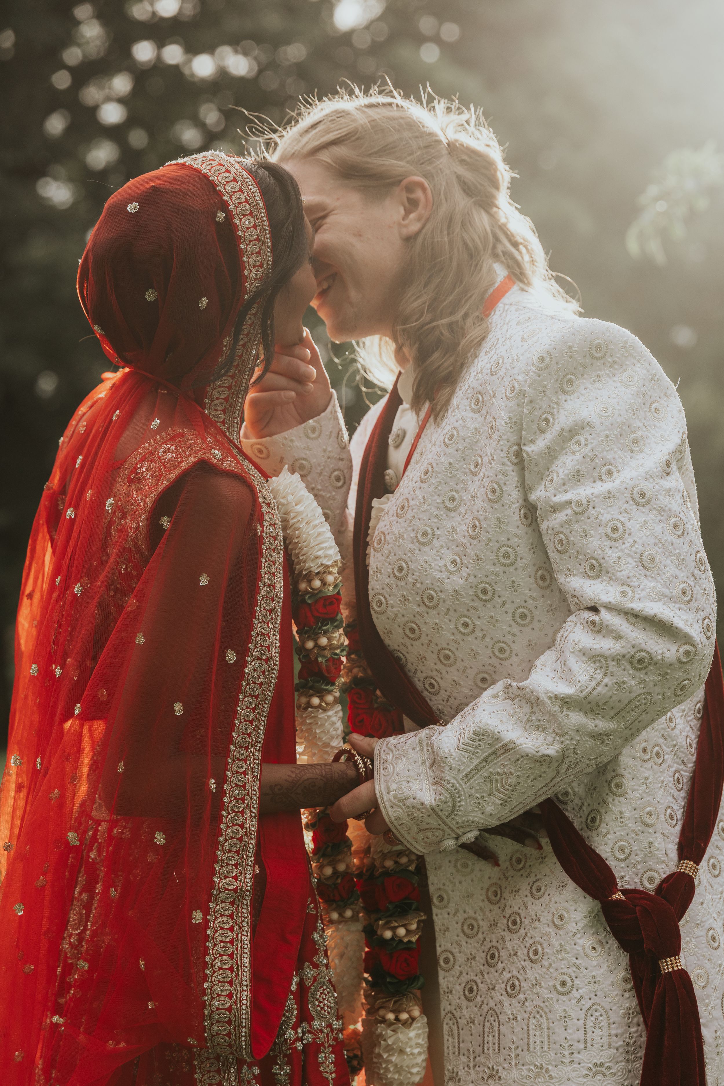 Two brides in traditional Indian wedding attire share an intimate moment, one in red lehenga and one in white sherwani.