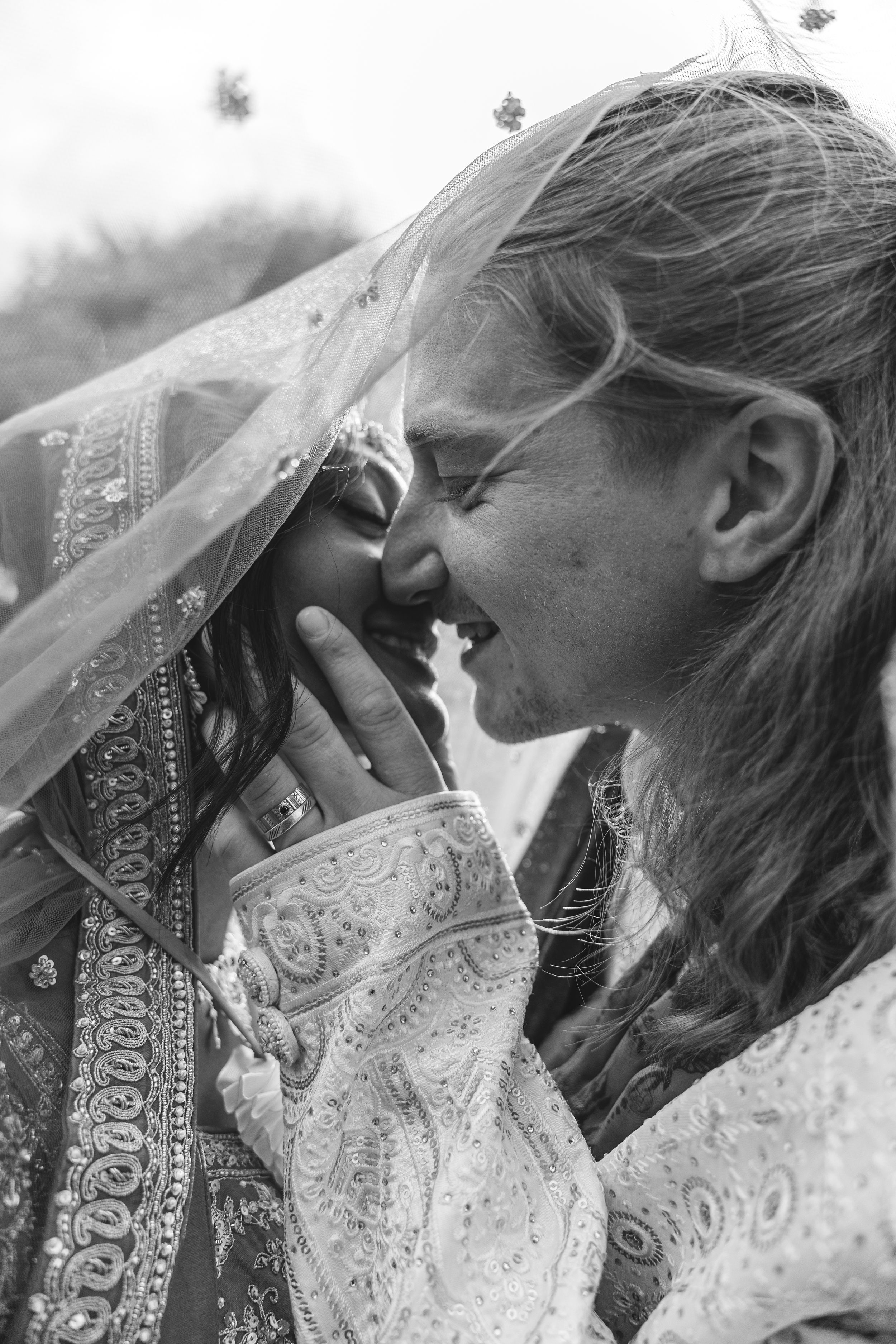 Black and white photo of two brides sharing a joyful moment, one in ornate Indian bridal attire with veil.