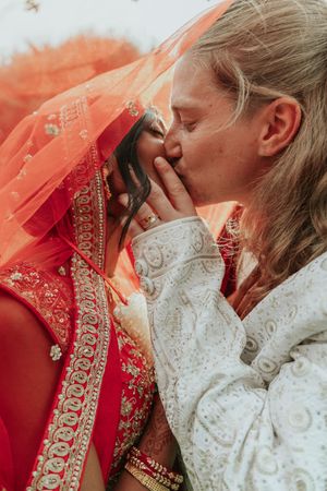 Couple sharing a romantic kiss, bride in red Indian bridal lehenga with veil, groom in white embroidered sherwani.