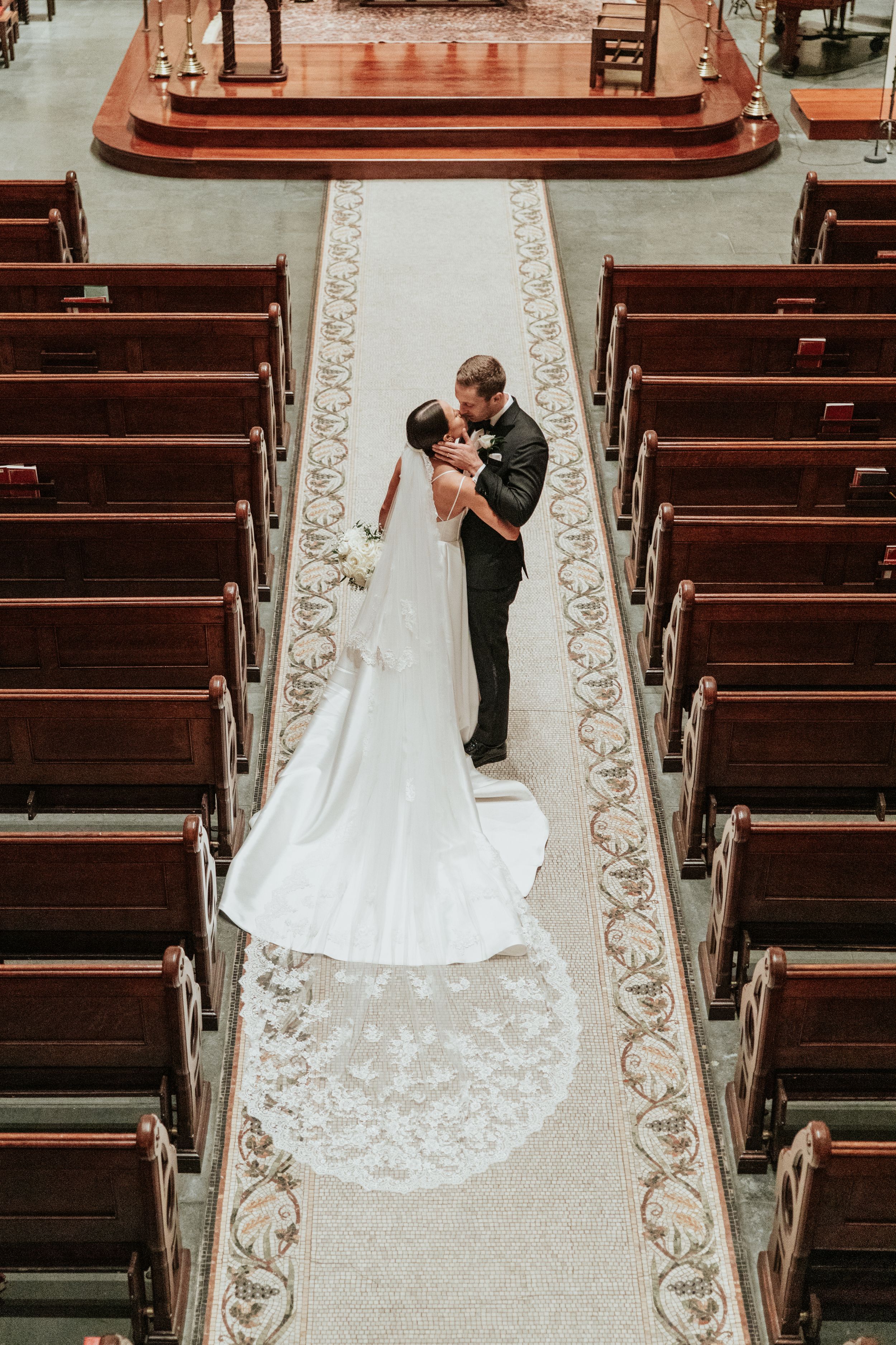 Bride and groom share a kiss at the altar of an elegant church, with ornate pews and a lace-trimmed cathedral veil.