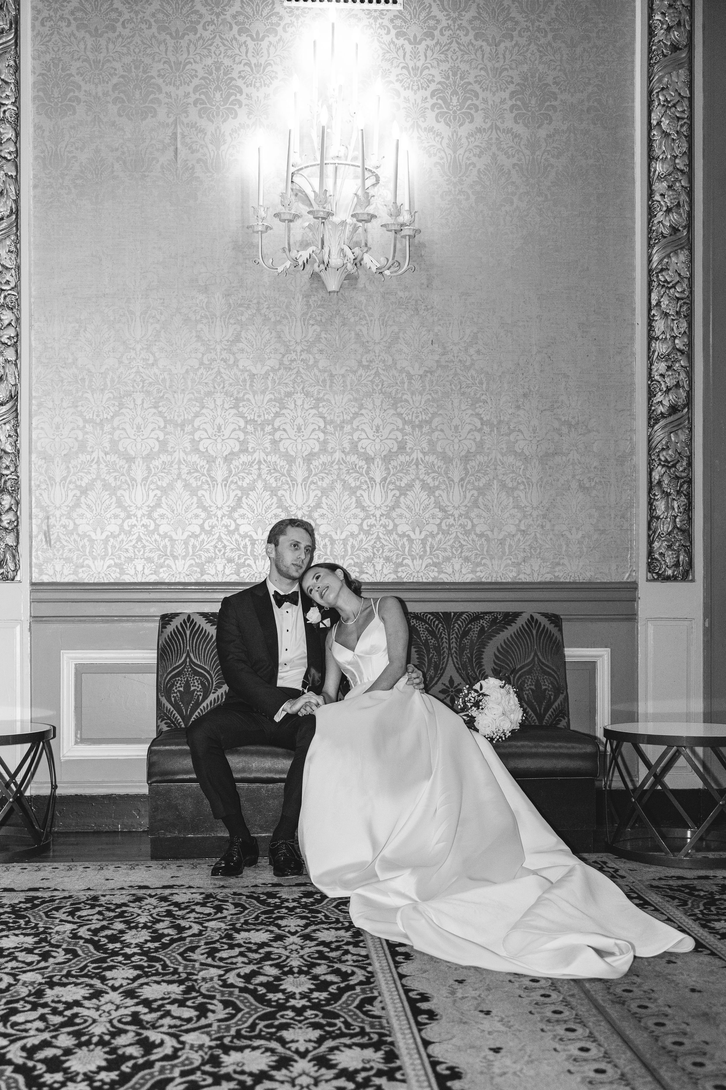 Black and white photo of bride and groom sitting on elegant sofa beneath chandelier in ornate ballroom.