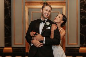 Bride and groom embracing and smiling in elegant black tie wedding attire inside a grand marble venue.