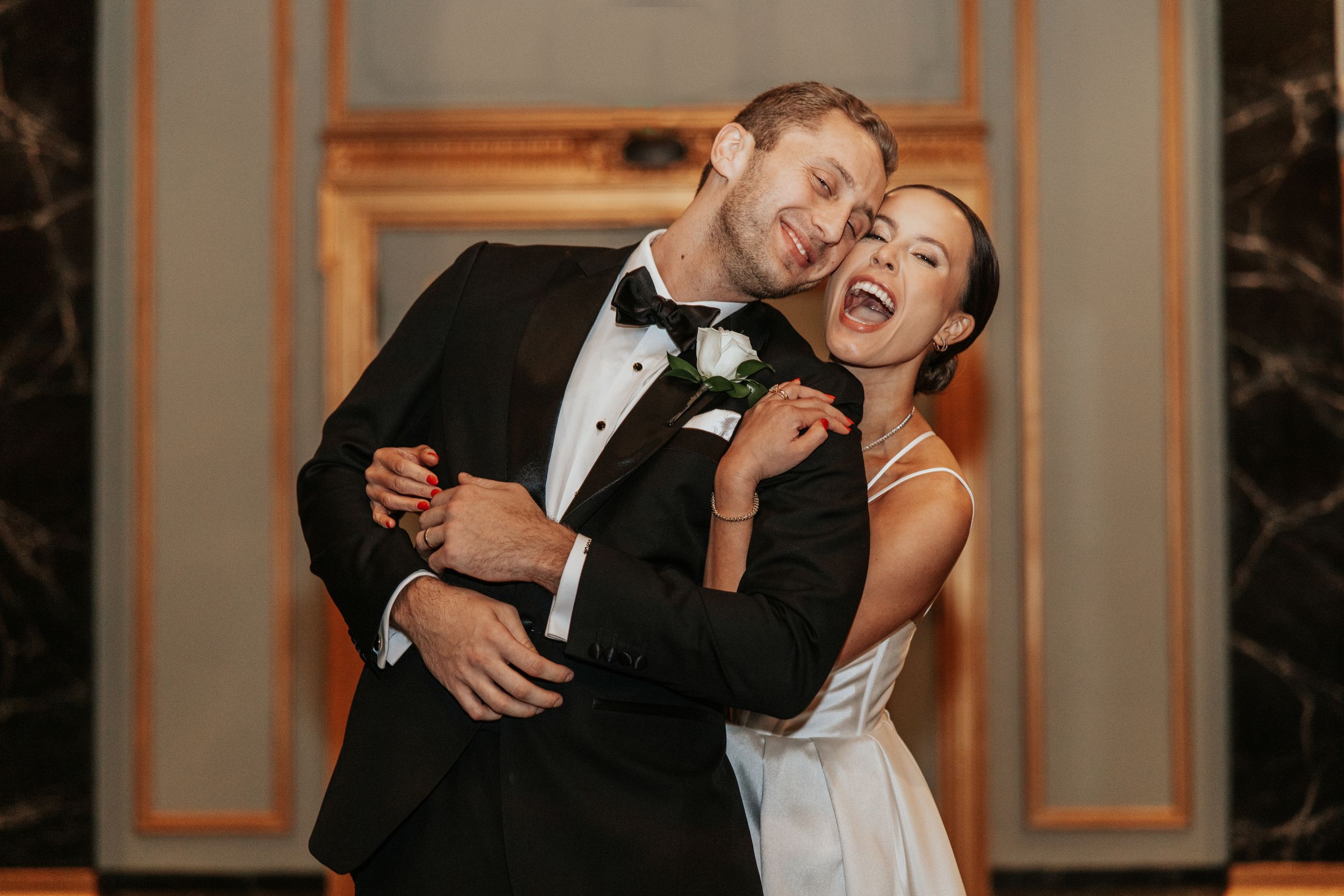 Joyful bride and groom embracing and laughing together in elegant wedding attire at an upscale indoor venue.