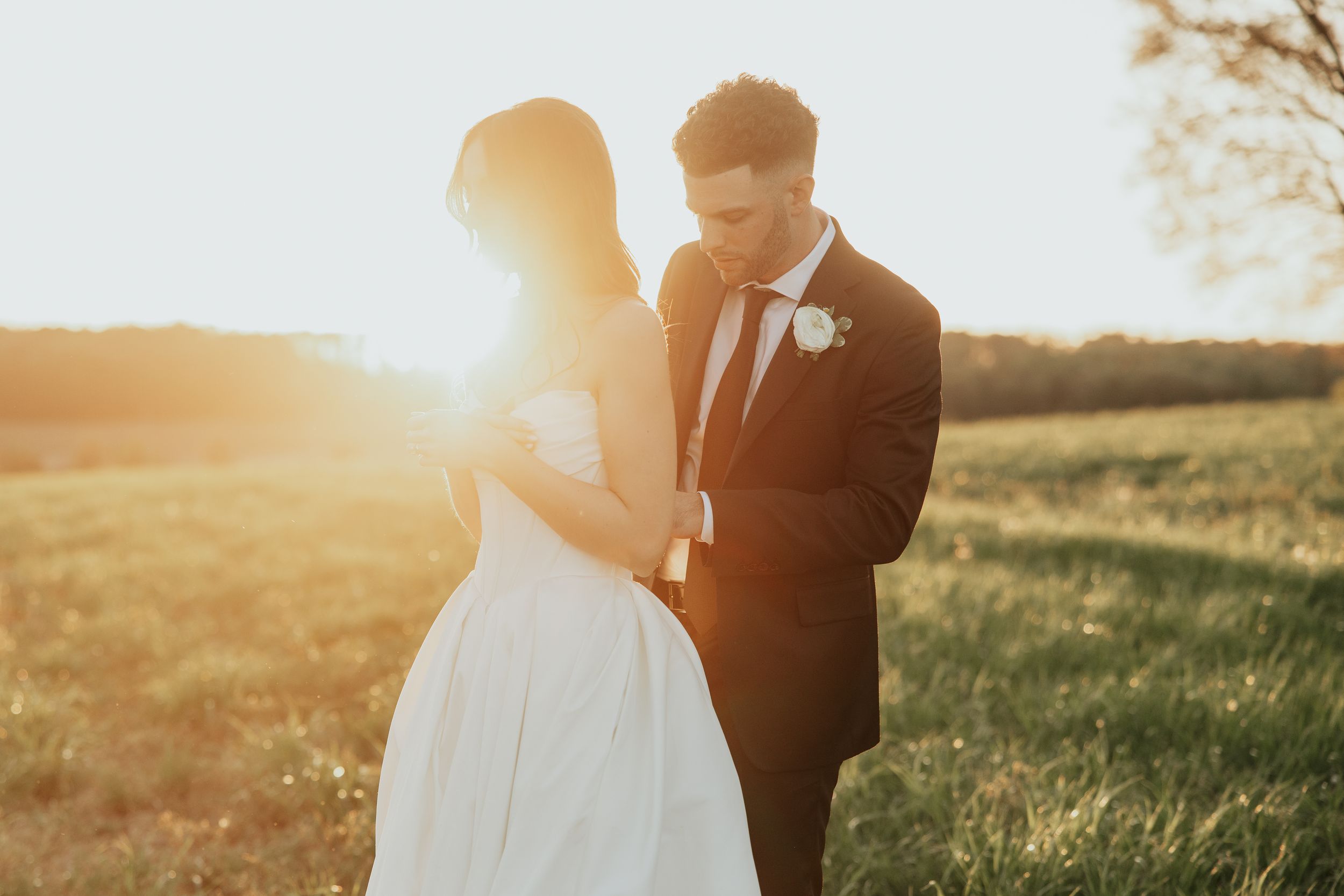 Couple sharing romantic golden hour moment in field, bride in white gown and groom in dark suit with boutonniere.