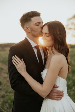 A groom in a black suit kisses his bride in a white strapless gown during a golden sunset outdoor wedding.