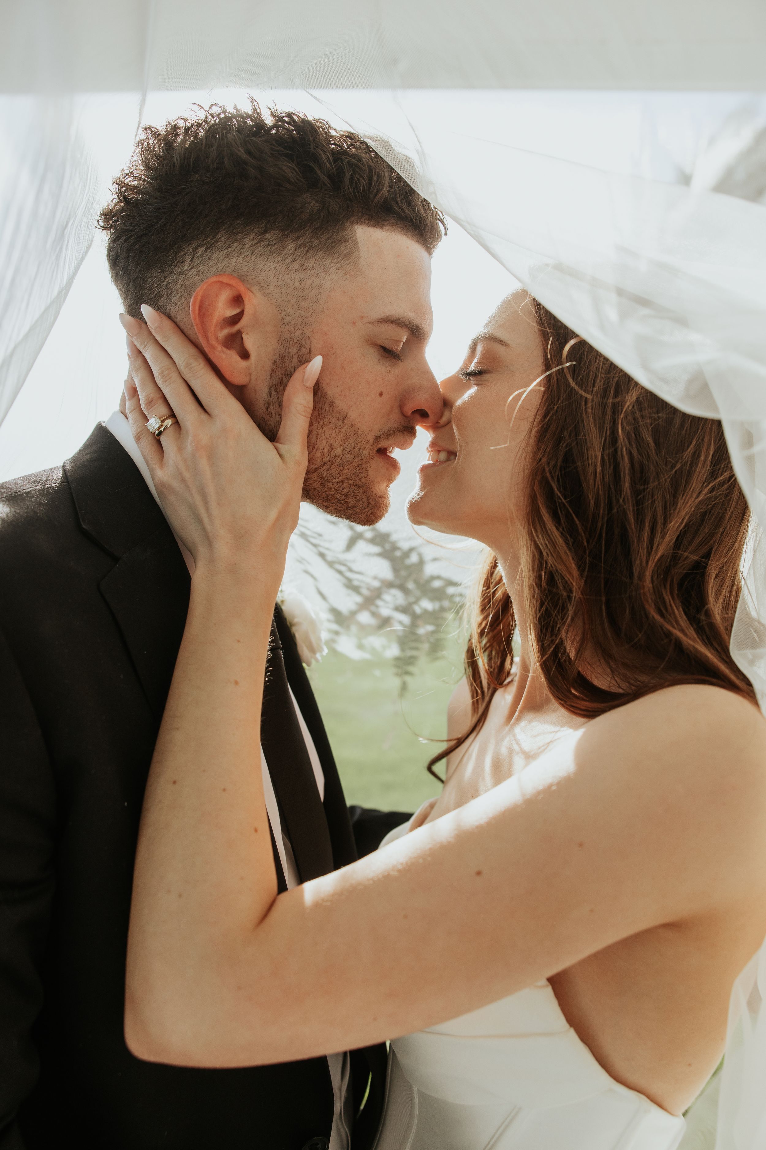 Bride and groom sharing a romantic almost-kiss under a wedding veil on their wedding day.