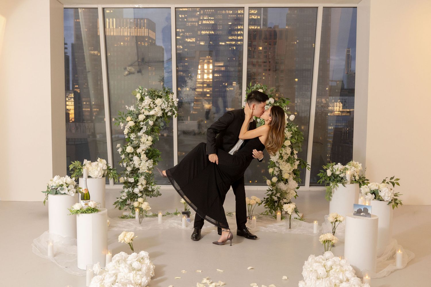 Couple sharing romantic dip kiss surrounded by white floral arrangements and city skyline views at elegant venue.